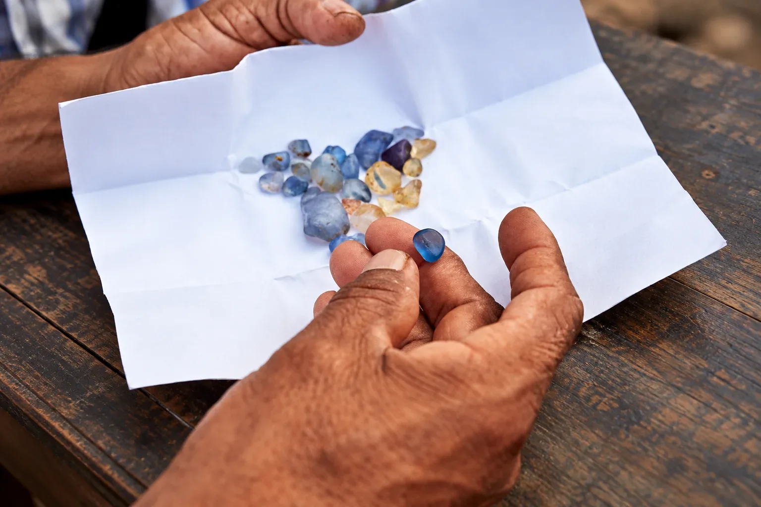 Hands examining a rough blue sapphire above a parcel of uncut stones