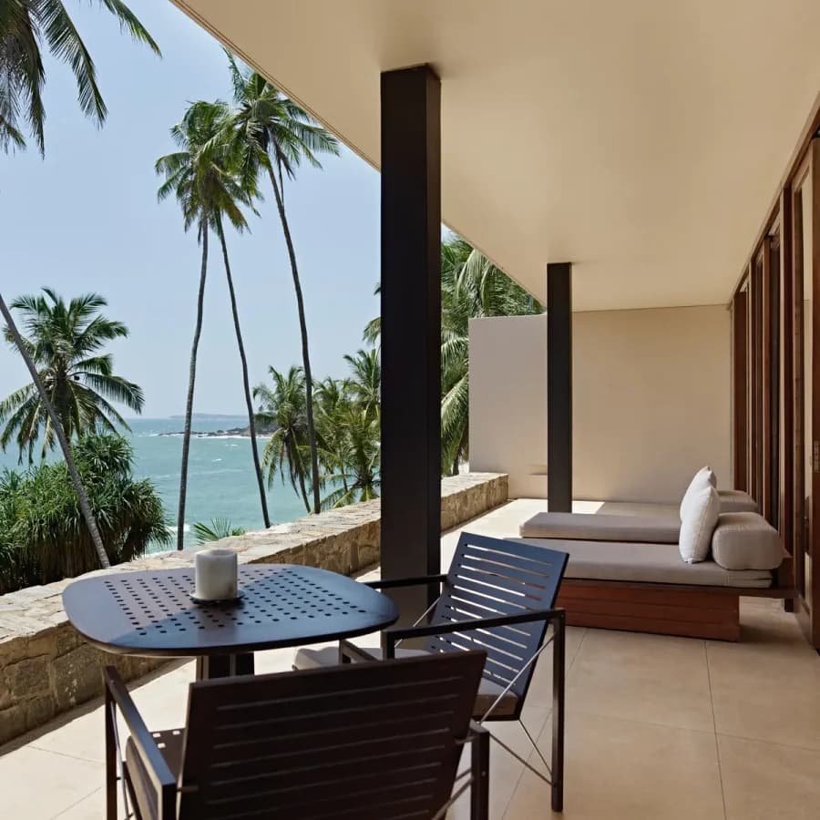 Amanwella suite ocean terrace with dining table, lounge chairs and sea views beyond palm trees.